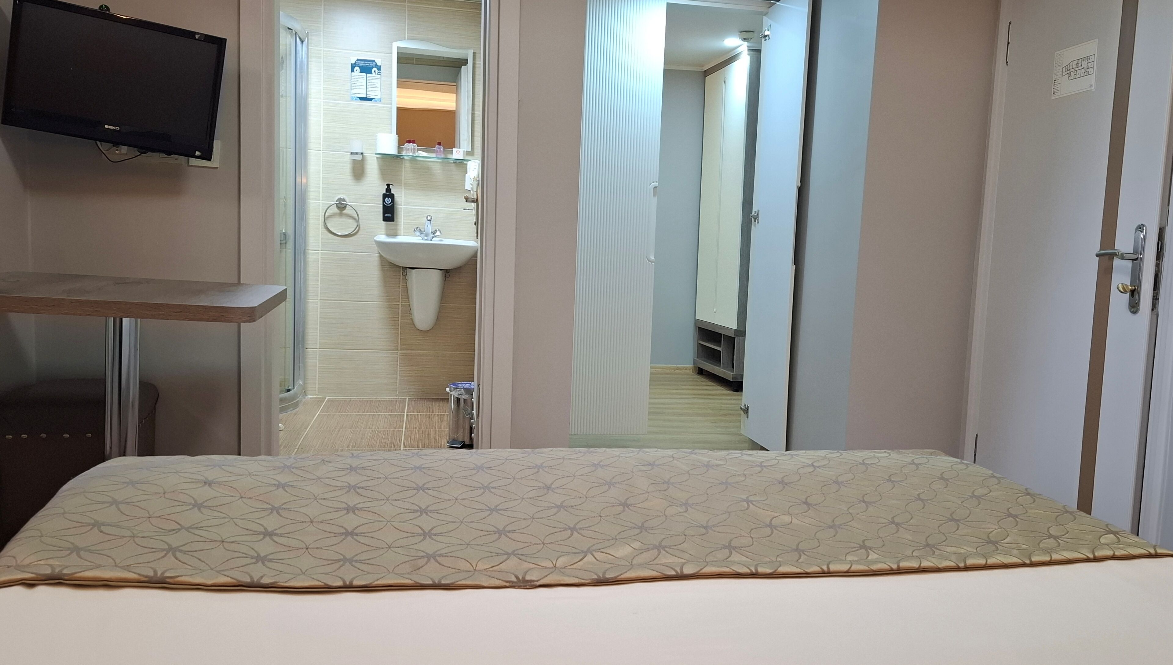A hotel room with a bed, a TV mounted on the wall, a bathroom with a sink and mirror, and a door leading to another room.