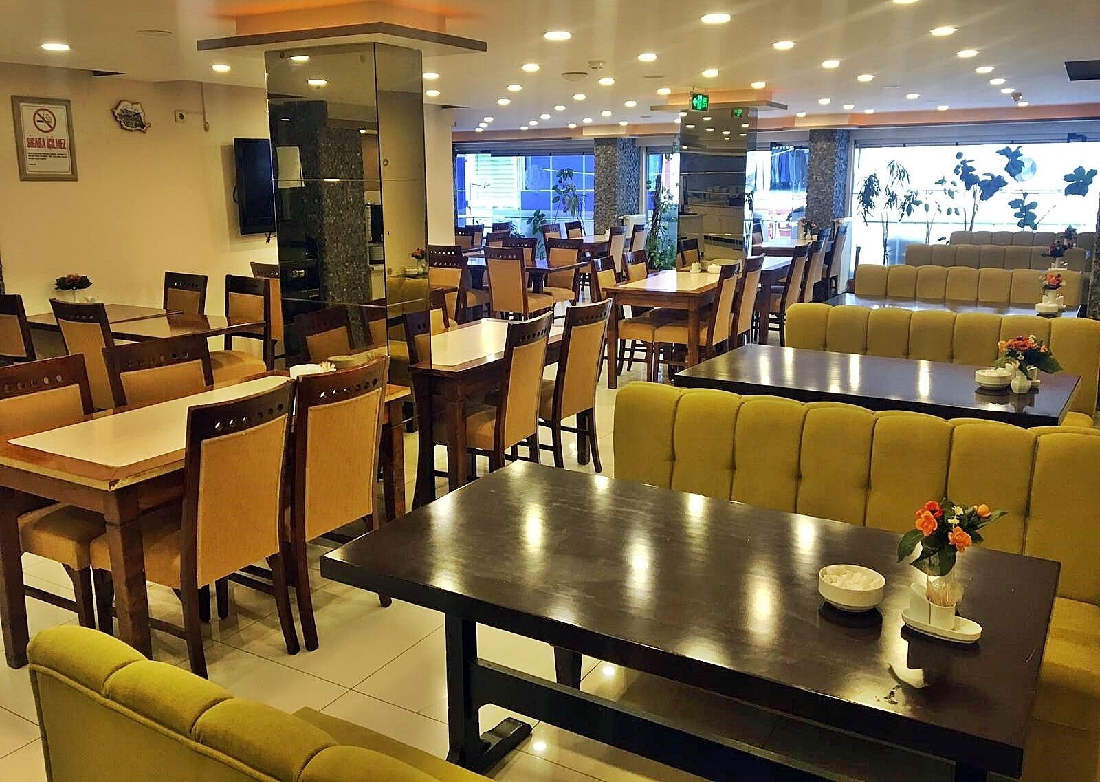 A restaurant with multiple dining tables and booths, a television mounted on the wall, and a large window with plants outside.