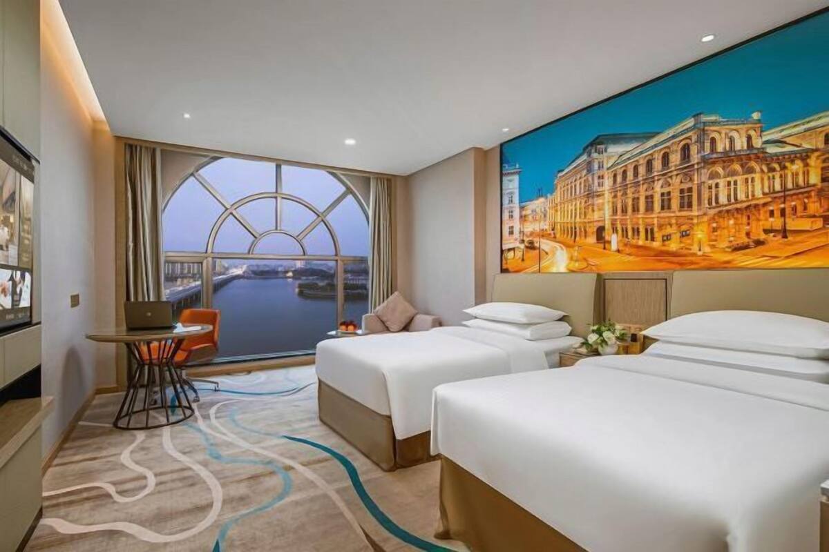 A hotel room with two beds, a desk, a laptop, and a large window with a view of a cityscape.
