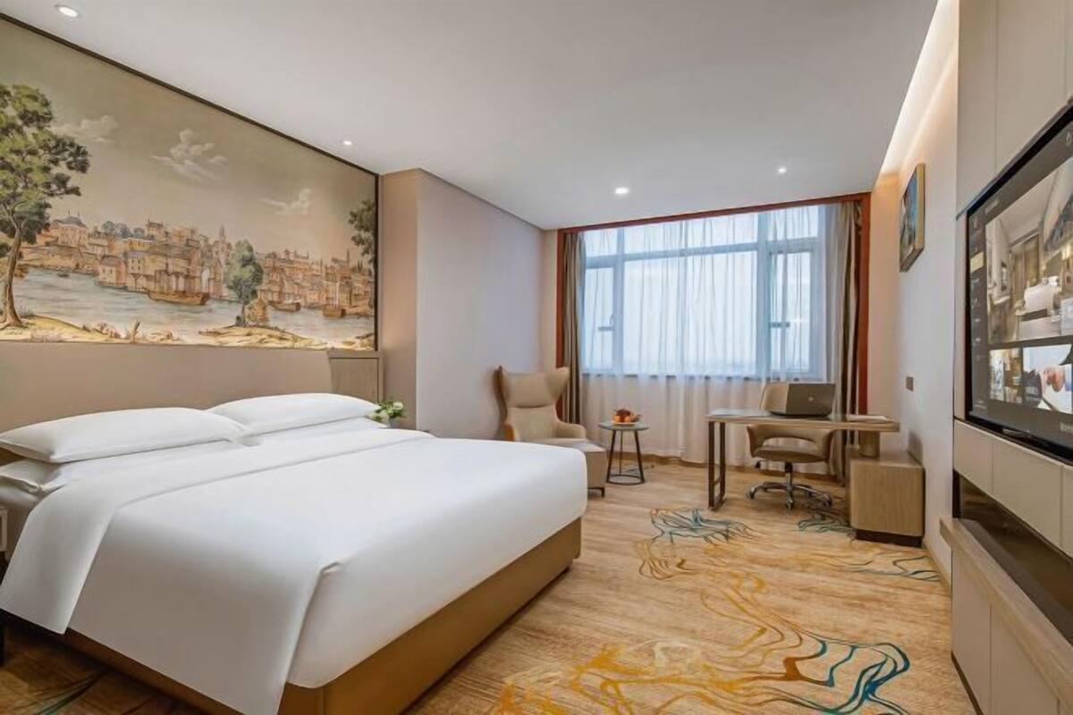 A hotel room with a large bed, a desk with a chair, a TV, and a mural on the wall.