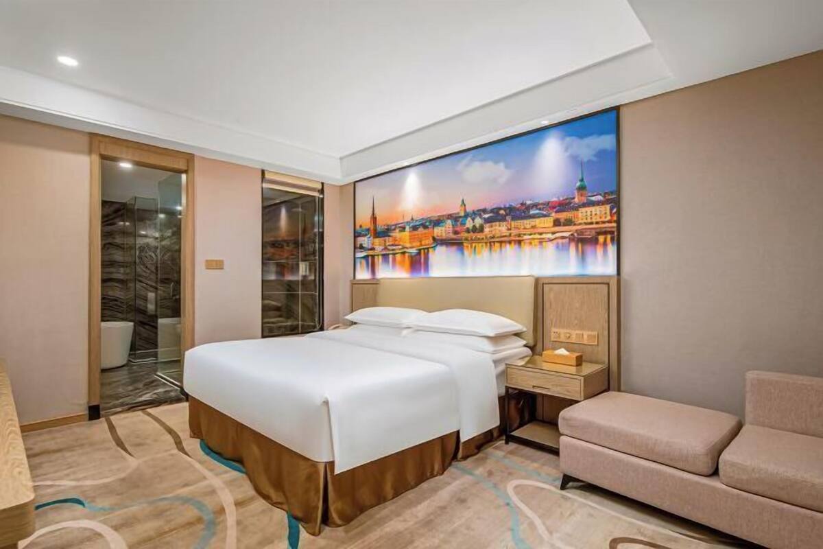 A hotel room with a bed, a nightstand, a chair, and a large cityscape painting on the wall.