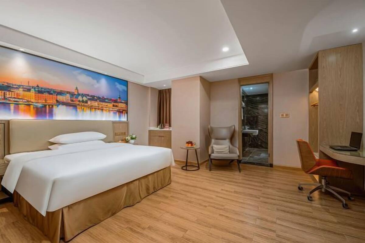 A hotel room with a bed, a desk, a chair, and a painting of a cityscape.