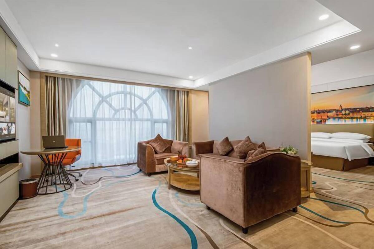 A modern hotel room with a large window, a sofa set, a desk with a laptop, and a bed with a view of the cityscape.