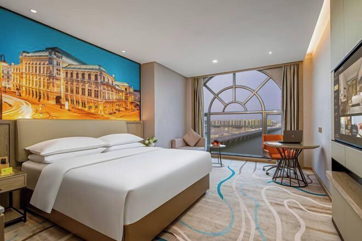 A hotel room with a large bed, a desk with a laptop, a window with a view of a cityscape, and a painting on the wall.