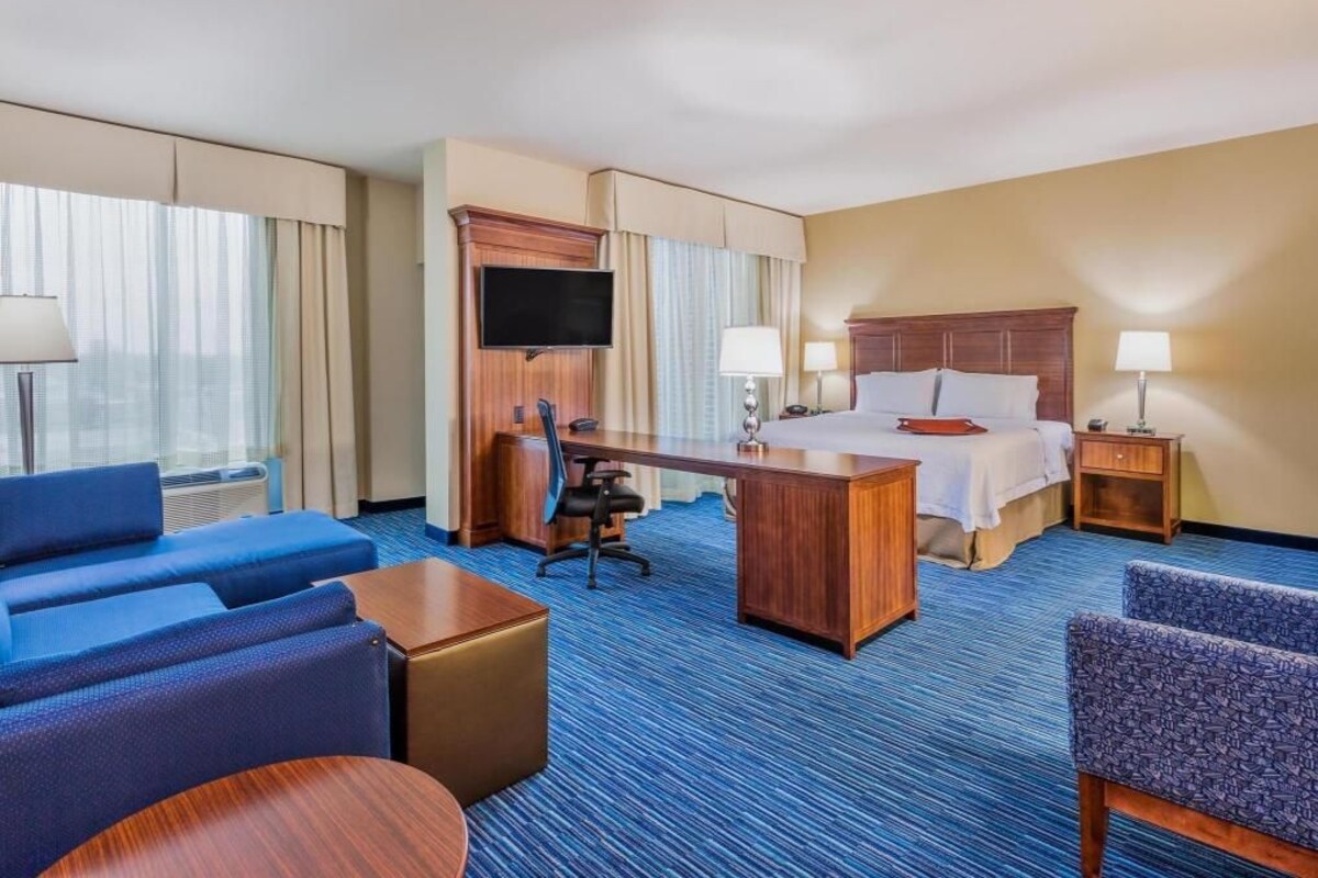 Habitación de hotel con cama, escritorio, dos sillas, televisor y ventana con cortinas.