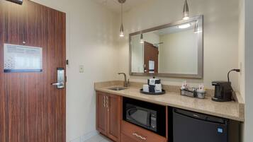 A modern bathroom with a wooden door, a sink, a microwave, a coffee maker, and a mirror.