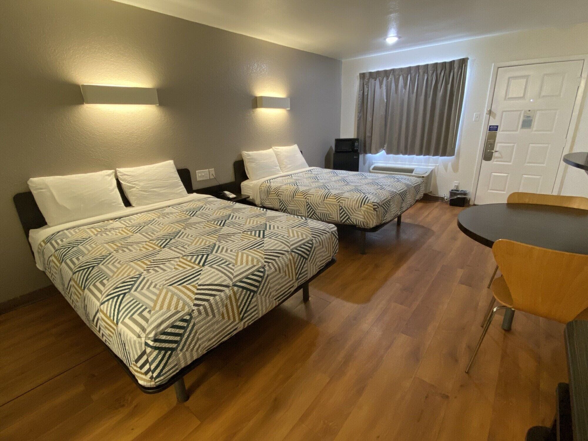 A hotel room with two beds, a small table, and a chair. There are wall-mounted lights and a window with curtains.
