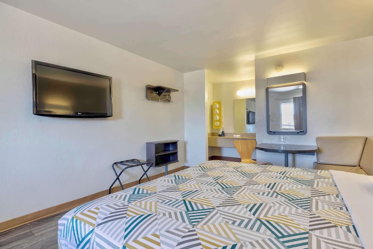 Ett hotellrum med en säng, en tv som är monterad på väggen, ett litet skrivbord, en stol, en spegel och en sminkbord med spegel och belysning.