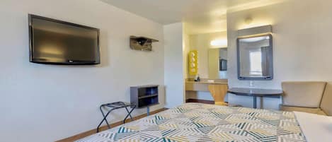 A hotel room with a bed, a TV mounted on the wall, a small desk, a chair, a mirror, and a vanity with a mirror and lights.