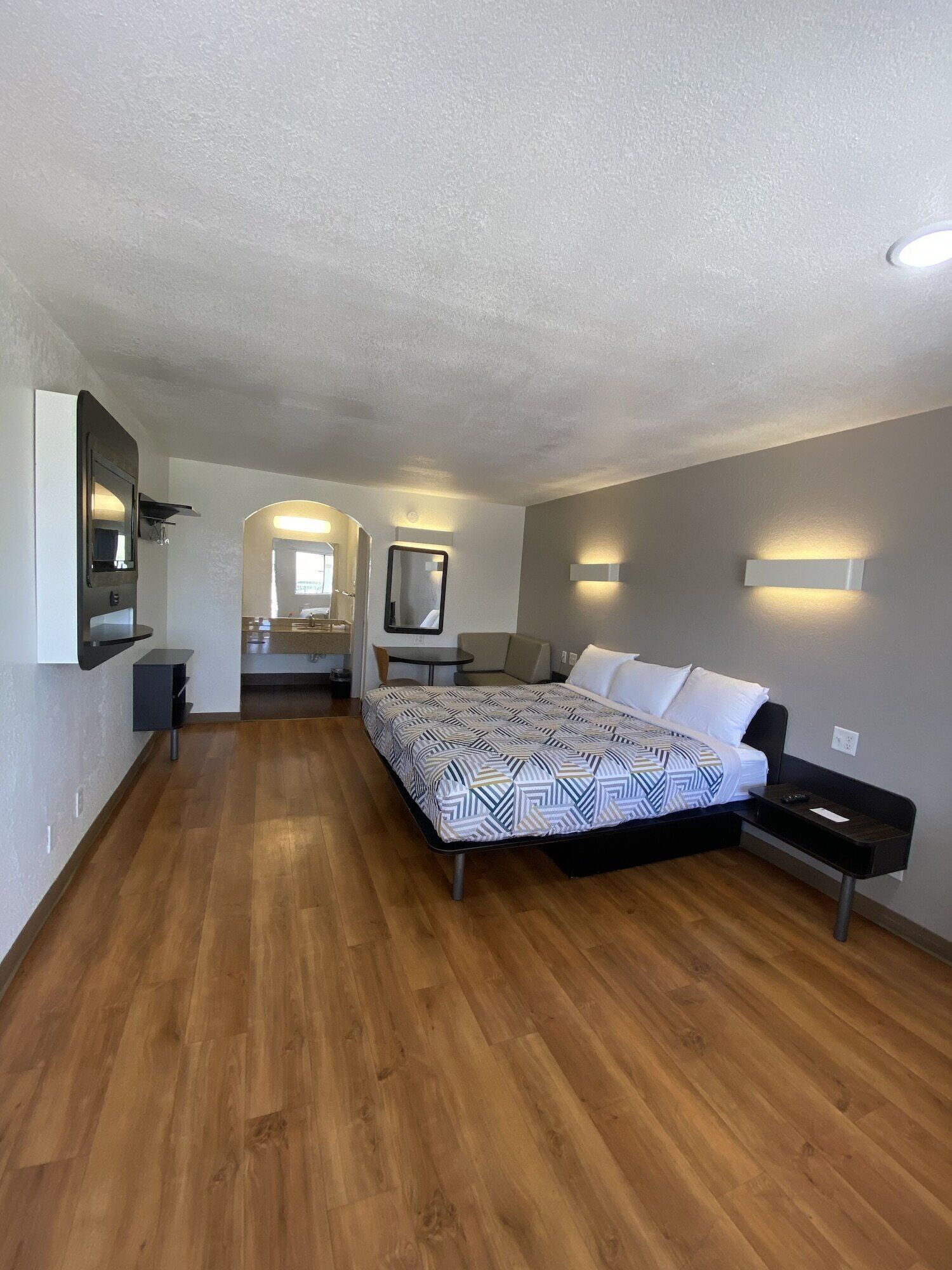 A modern hotel room with a wooden floor, a large bed with patterned bedding, a small desk, a chair, a mirror, and a wall-mounted light fixture.