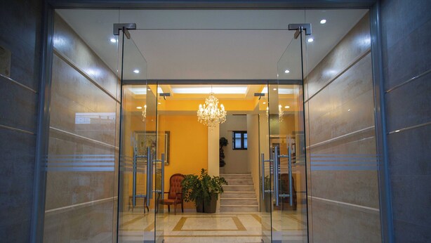 A modern lobby with a glass entrance, a chandelier, a staircase, and a seating area with a plant. - Hotel Giorgio (Acharnes)