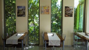 A dining area with tables and chairs, large windows with a view of greenery, and artwork on the walls. - Hotel Giorgio (Acharnes)