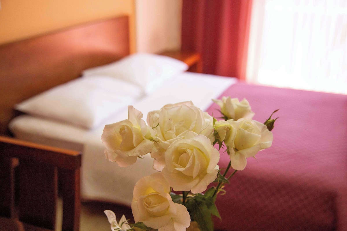 A bed with white linens and a pink bedspread, a wooden headboard, and a vase with white flowers in the foreground.
