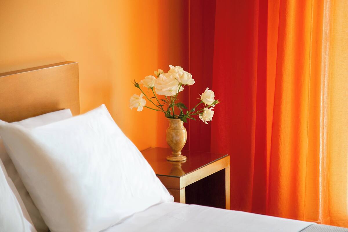 A hotel room with a bed, white pillows, a vase with white flowers, and a red curtain.