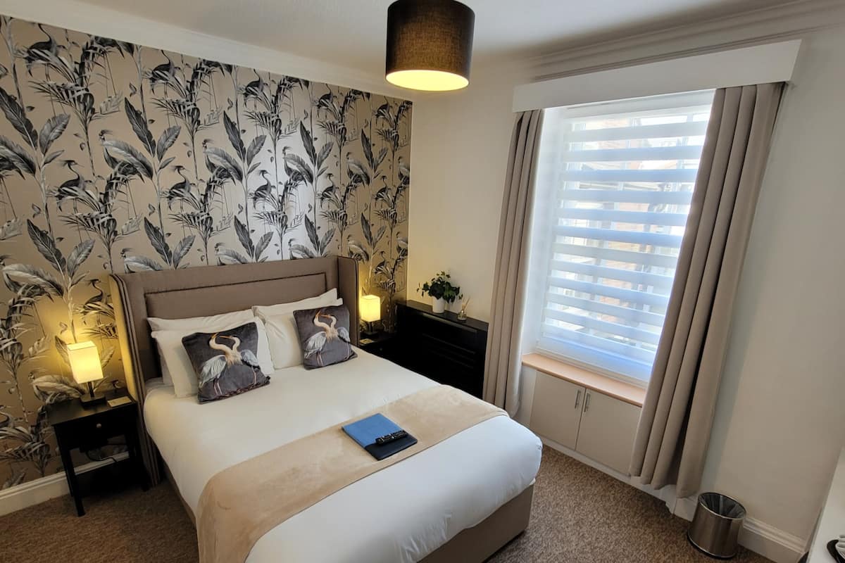 Un dormitorio con una cama grande, una mesita de noche con lámpara, una ventana con persianas, y una pared con estampado floral.