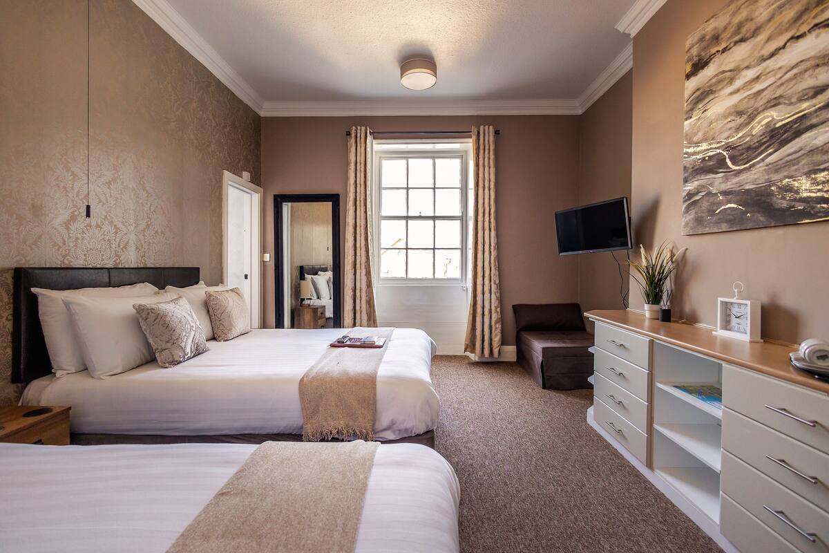 Habitación de hotel con dos camas, un escritorio, televisor y un ventanal con cortinas.