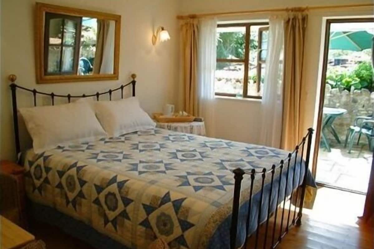 A bedroom with a bed, a nightstand, a window with curtains, and a view of the outdoors.