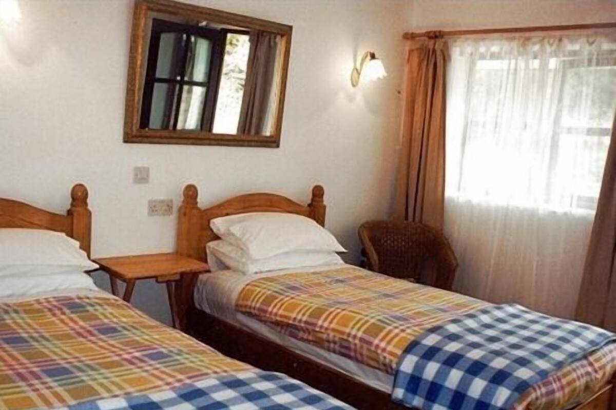 A room with two wooden beds, each with a checkered bedspread, a small table, a chair, a mirror, and a window with curtains.