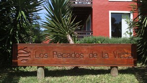 Ein hölzernes Schild mit dem Schriftzug "Los Pecados de la Viuda" vor einem Gebäude mit Balkonen und Fenstern.