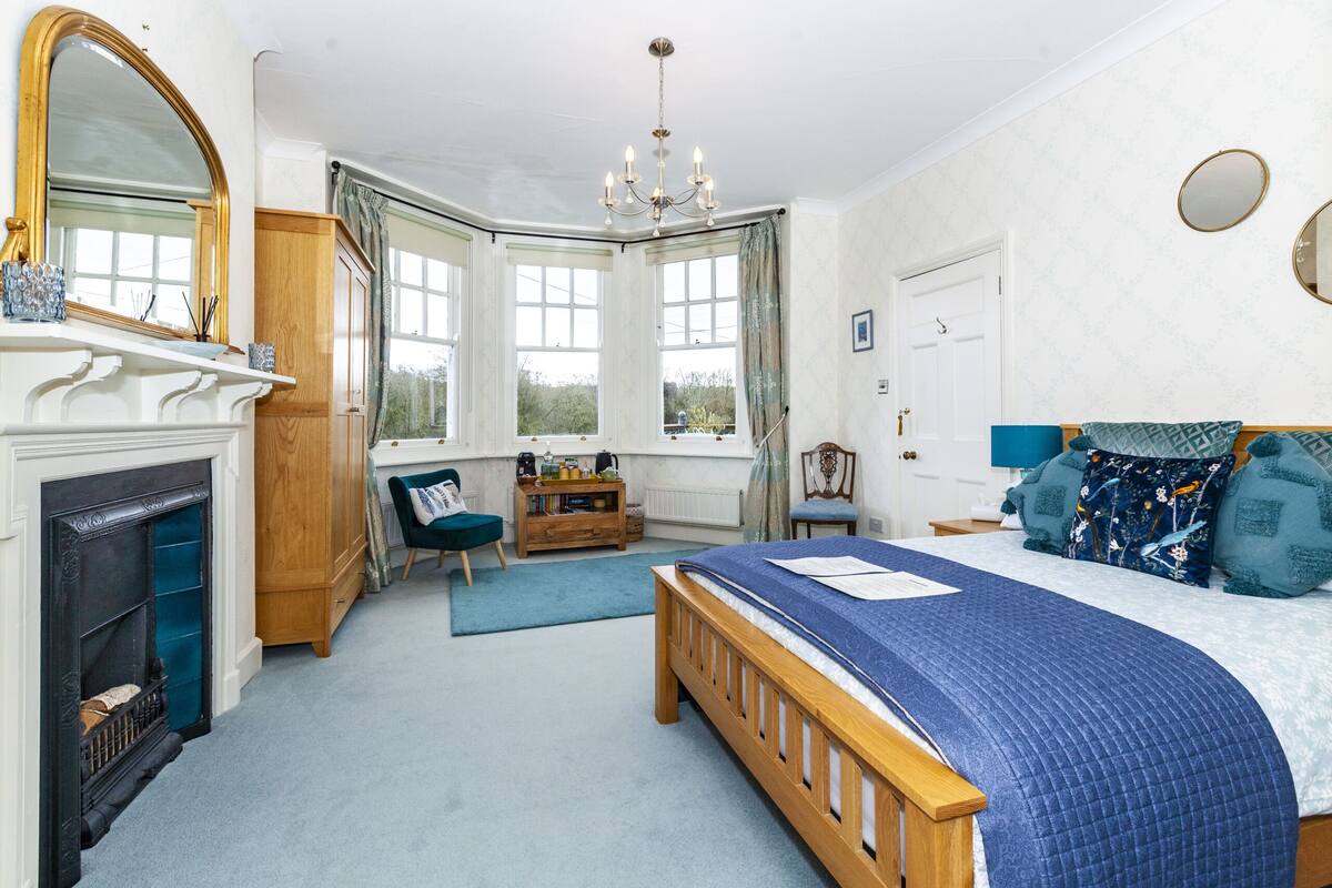 Een slaapkamer met een groot bed, een open haard, een houten kast, een bureau en een raam met gordijnen.