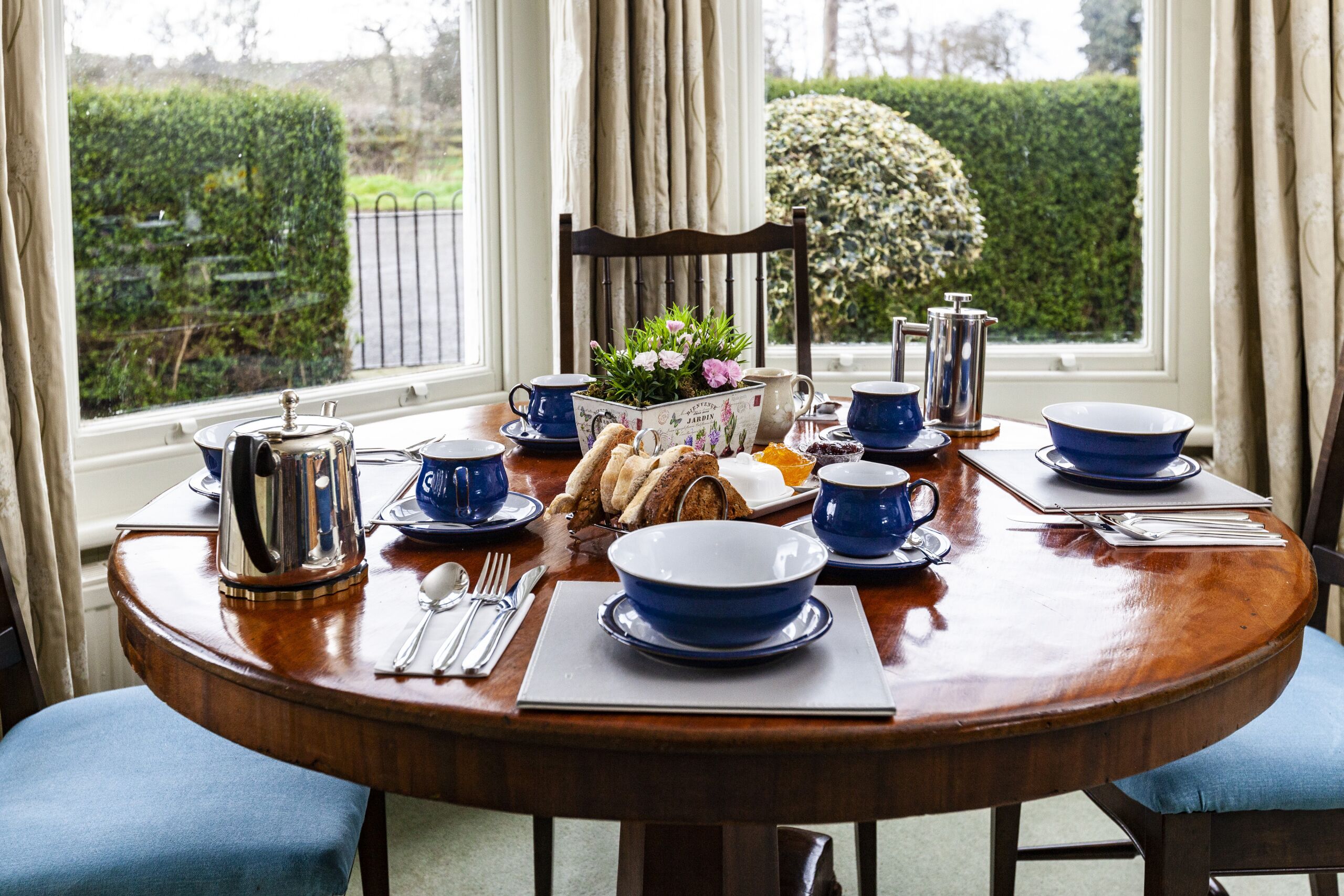Una mesa de madera redonda preparada para el desayuno, con vajilla azul y blanca, cubiertos y una cafetera.
