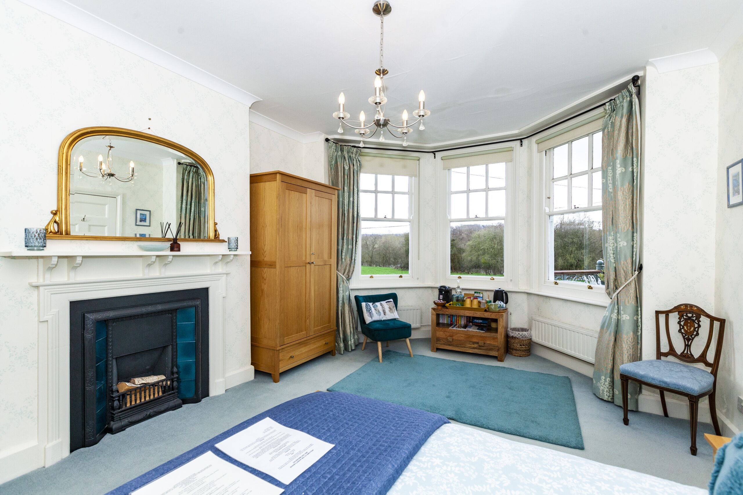 A bedroom with a fireplace, a large mirror, a chandelier, a wooden wardrobe, a chair, a bedside table, and a window with curtains.