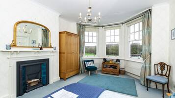 A bedroom with a fireplace, a large mirror, a chandelier, a wooden wardrobe, a chair, a bedside table, and a window with curtains.