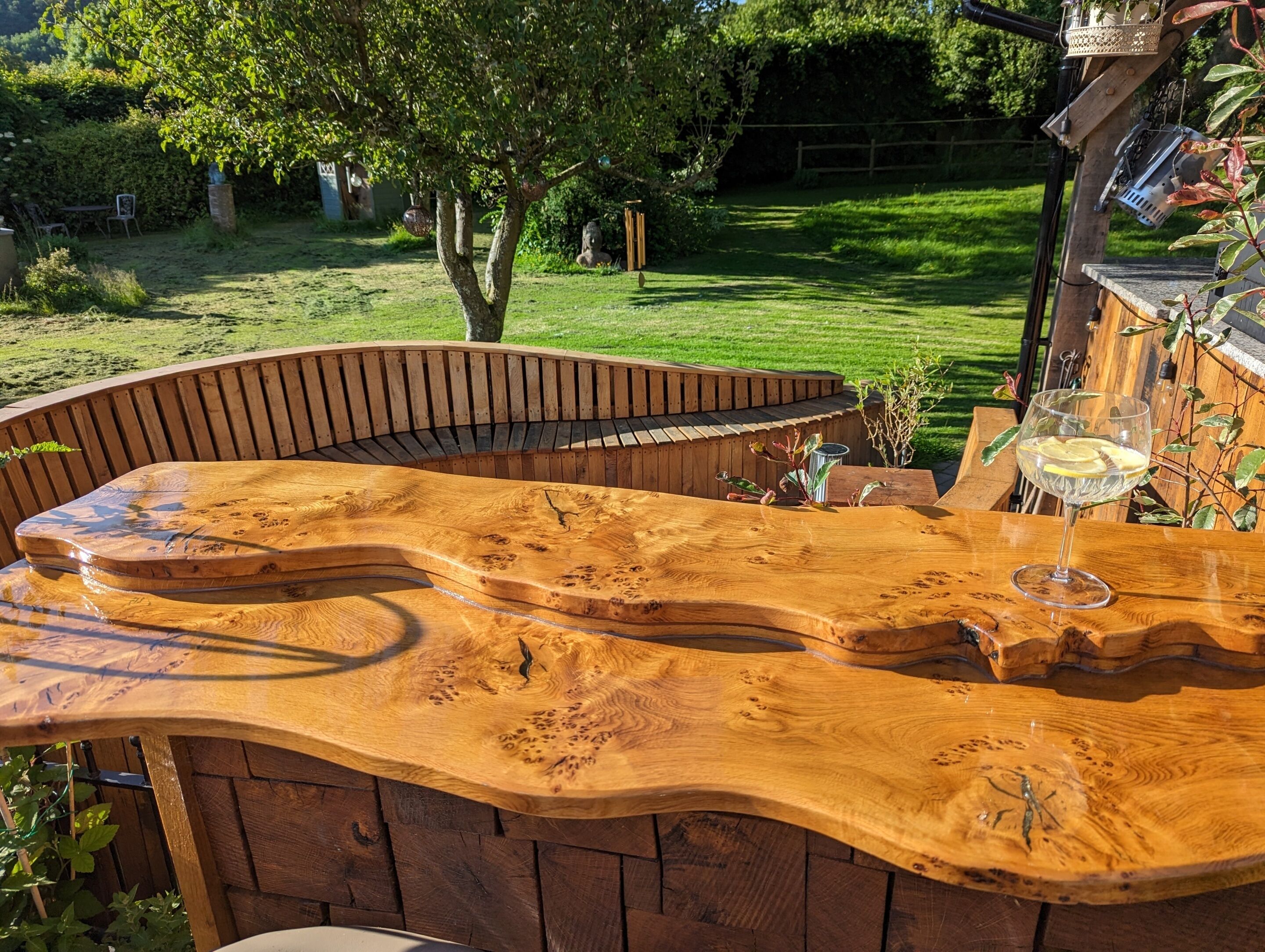 A wooden outdoor seating area with a curved bench and a glass table.
