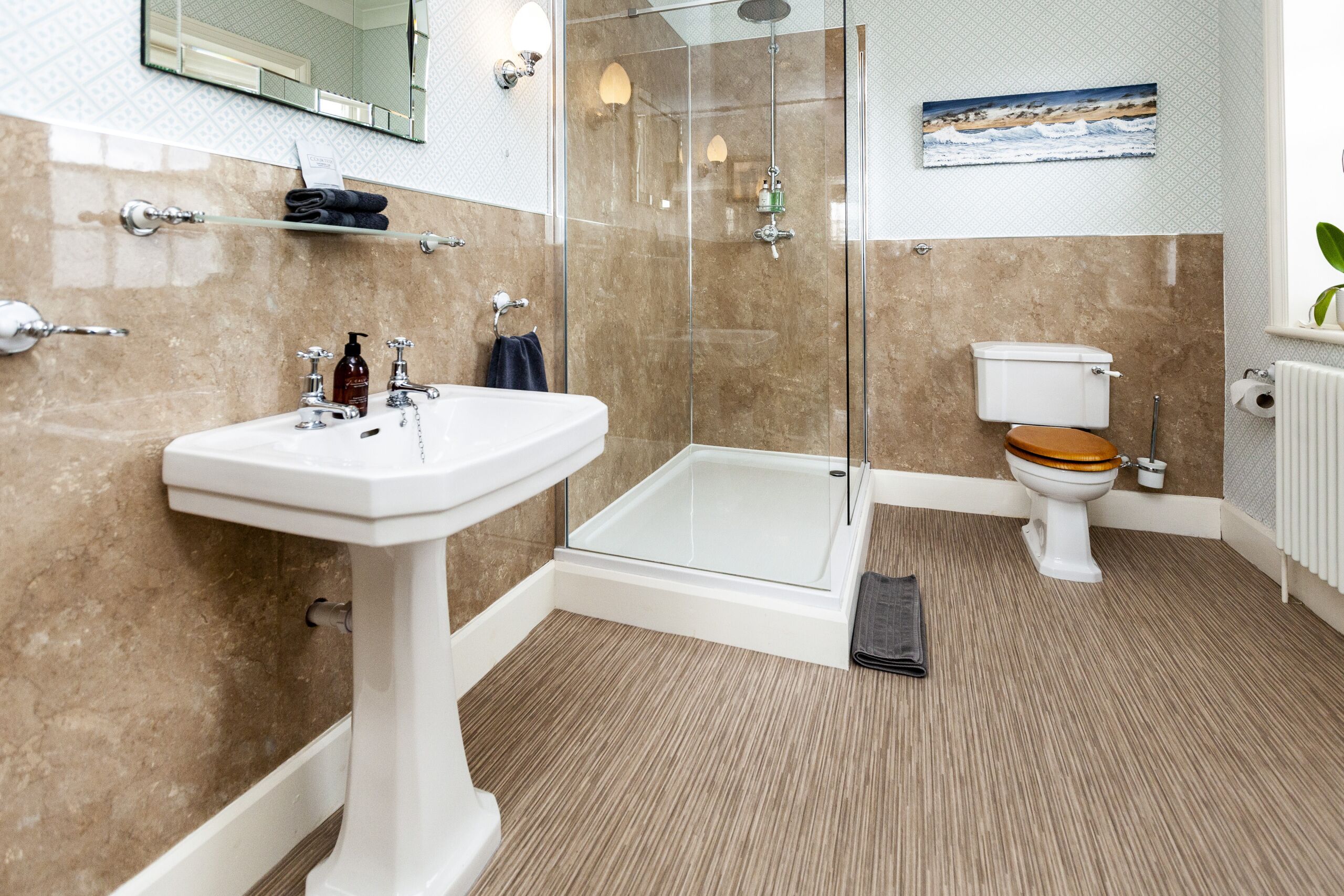 A modern bathroom with a glass-enclosed shower, a pedestal sink, and a toilet with a wooden seat.