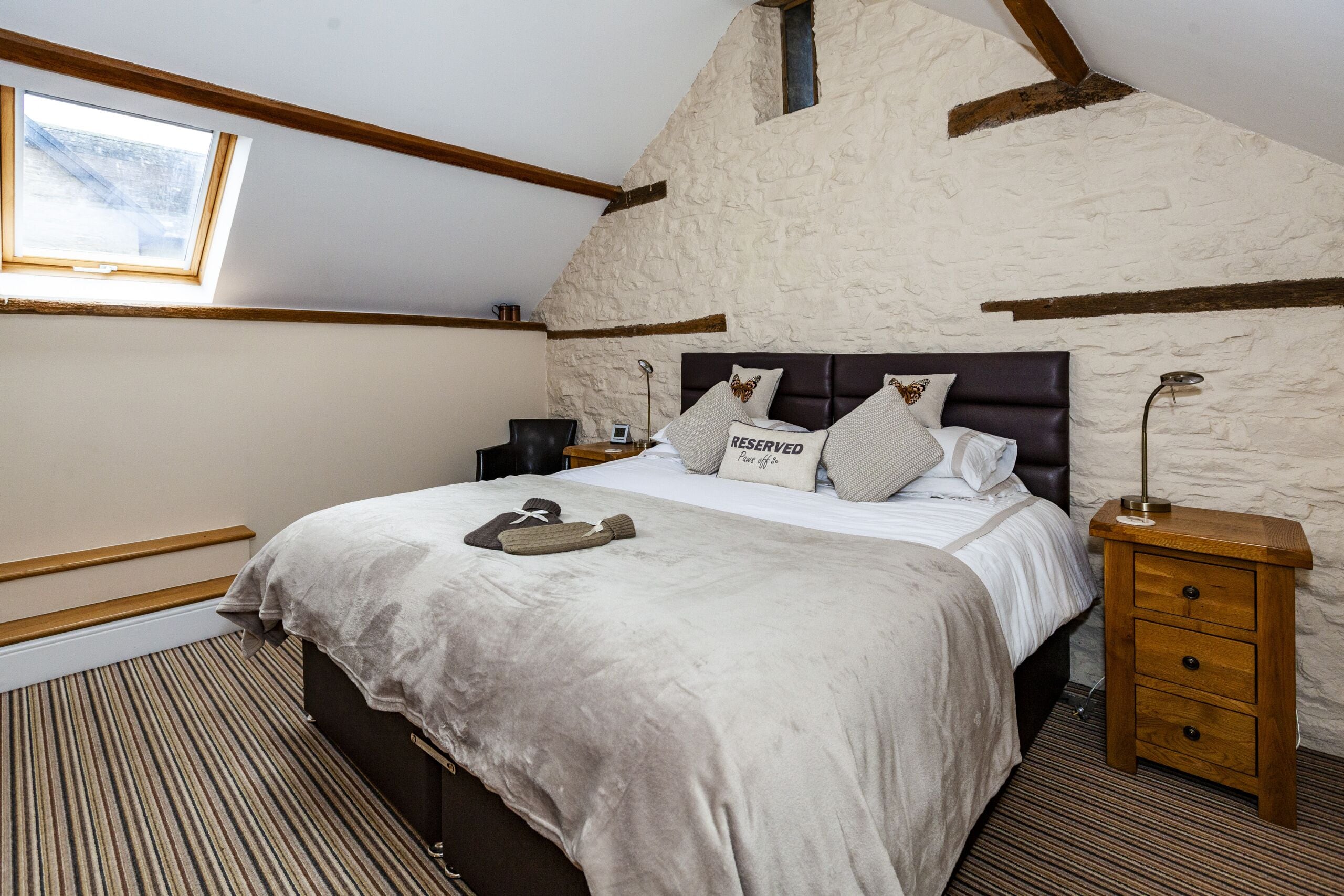 Quarto com teto inclinado, uma cama com cabeceira, mesa de cabeceira, abajur e janela.