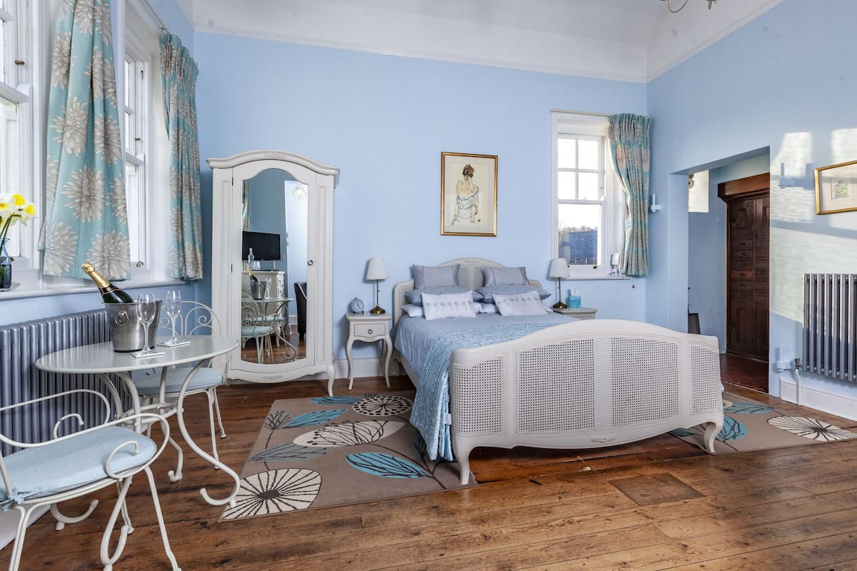 Een slaapkamer met een wit bed, een dressoir, een spiegel en een raam met gordijnen.