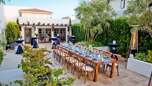 A long wooden table set for dining outdoors with chairs, potted plants, and a building in the background. - Hotel Marisol Coronado (Coronado)