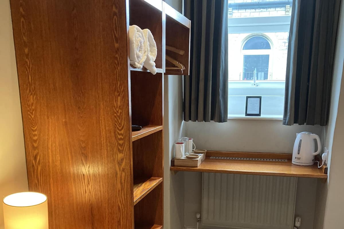 A hotel room with a wooden wardrobe, a bedside table with a lamp, a window with curtains, a radiator, and a bed.