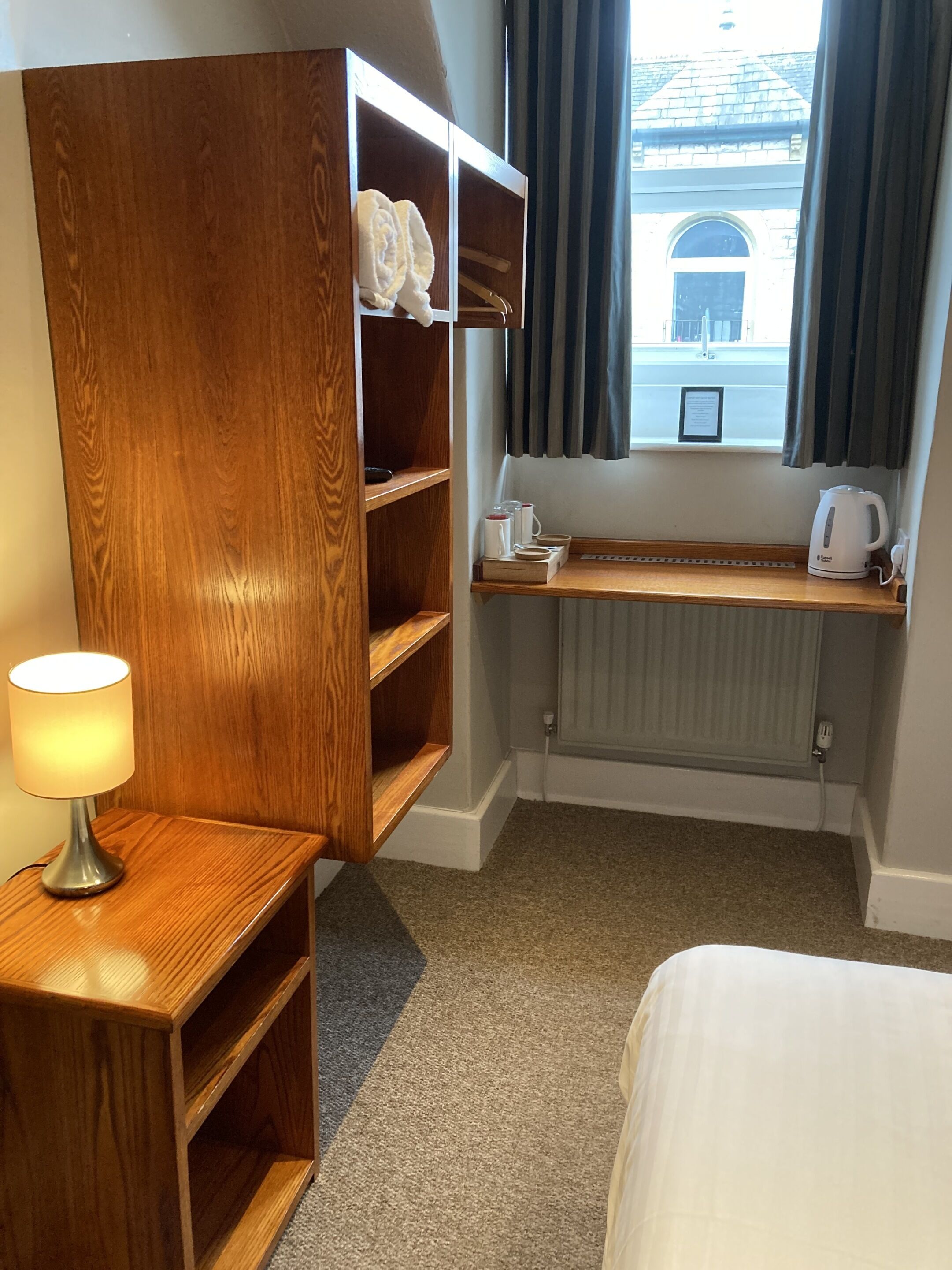 A hotel room with a wooden wardrobe, a bedside table with a lamp, a window with curtains, and a radiator.