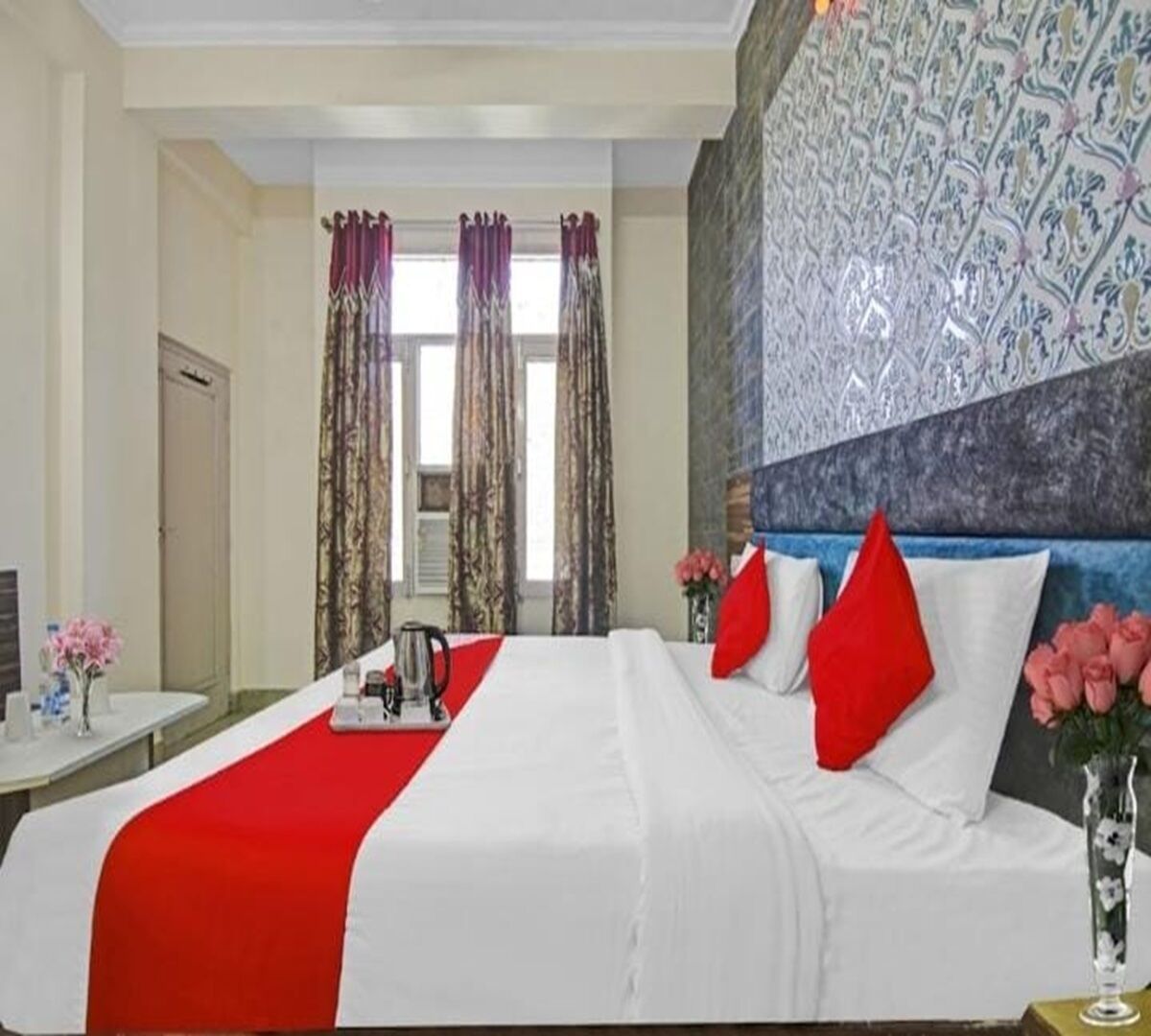 A hotel room with a large bed, red and white bedding, a coffee table with a kettle, and a decorative wall.