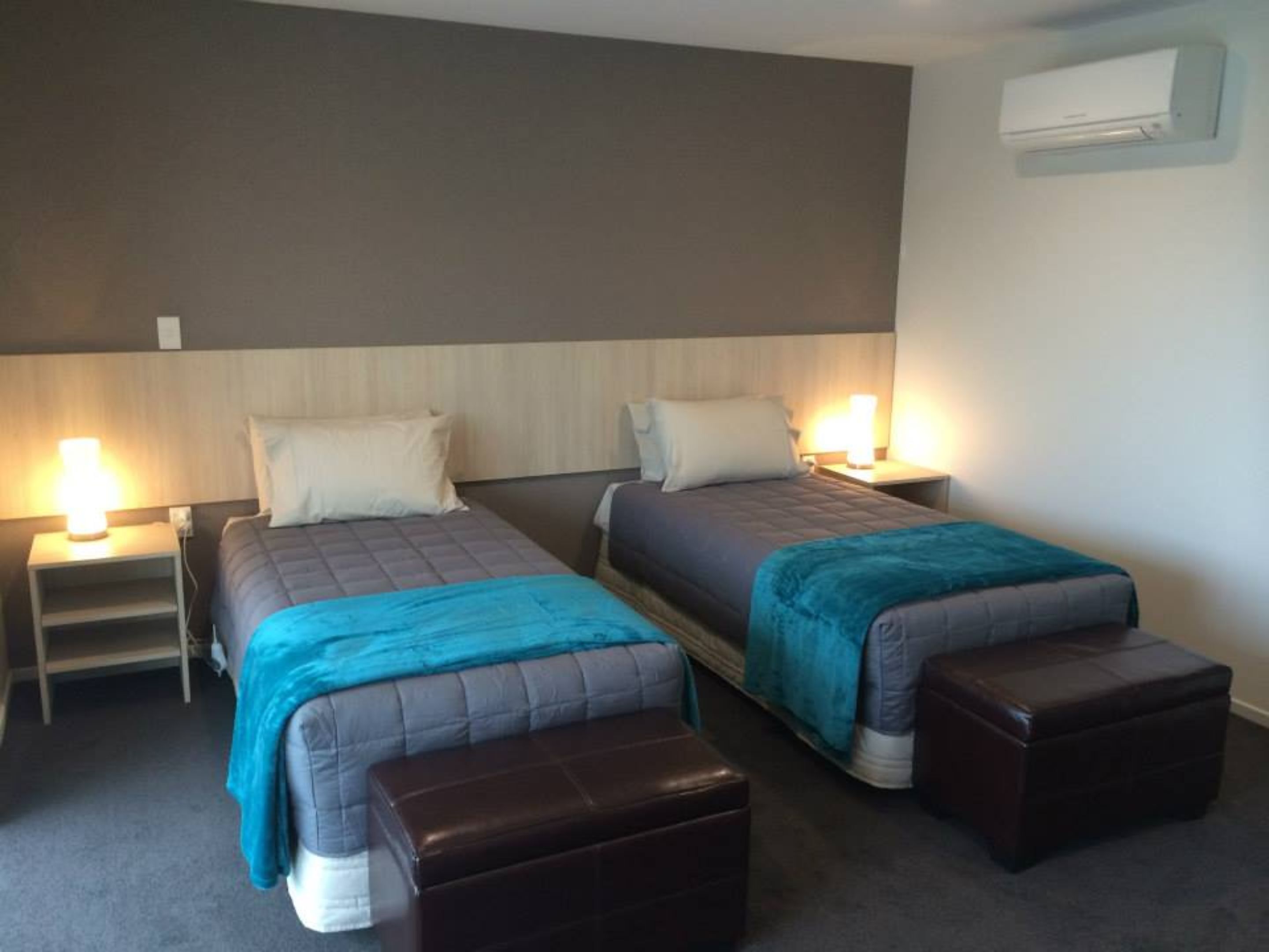 Una habitación de hotel con dos camas, cada una con cabecero, mesitas de noche con lámparas y un otoman marrón.