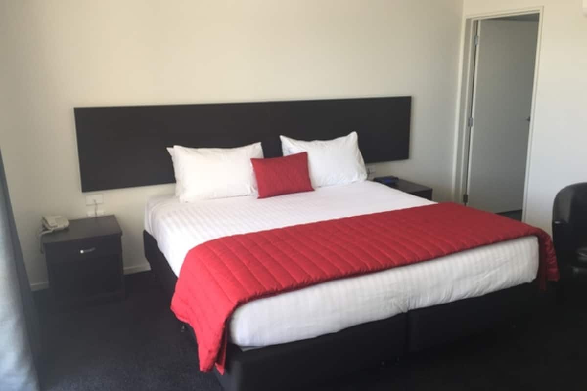 A hotel room with a large bed, white bedding, a red accent pillow, a black headboard, a nightstand with a phone, and a door.