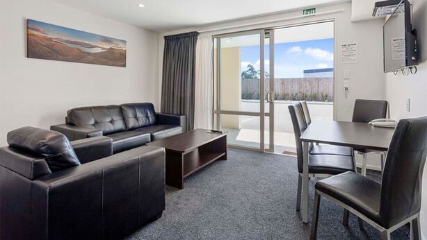 Una habitación de hotel moderna con un sofá de cuero negro, una mesita de centro, una mesa de comedor con sillas y un amplio ventanal con vistas al exterior.