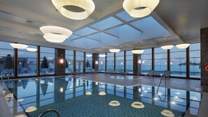 Ein großes Hallenbad mit klaren Glaswänden, Liegestühlen und modernen Beleuchtungselementen.