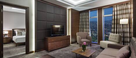 Quarto de hotel com cama, televisão sobre um armário de madeira, sofá, mesa de centro e vista para a cidade através de grandes janelas.