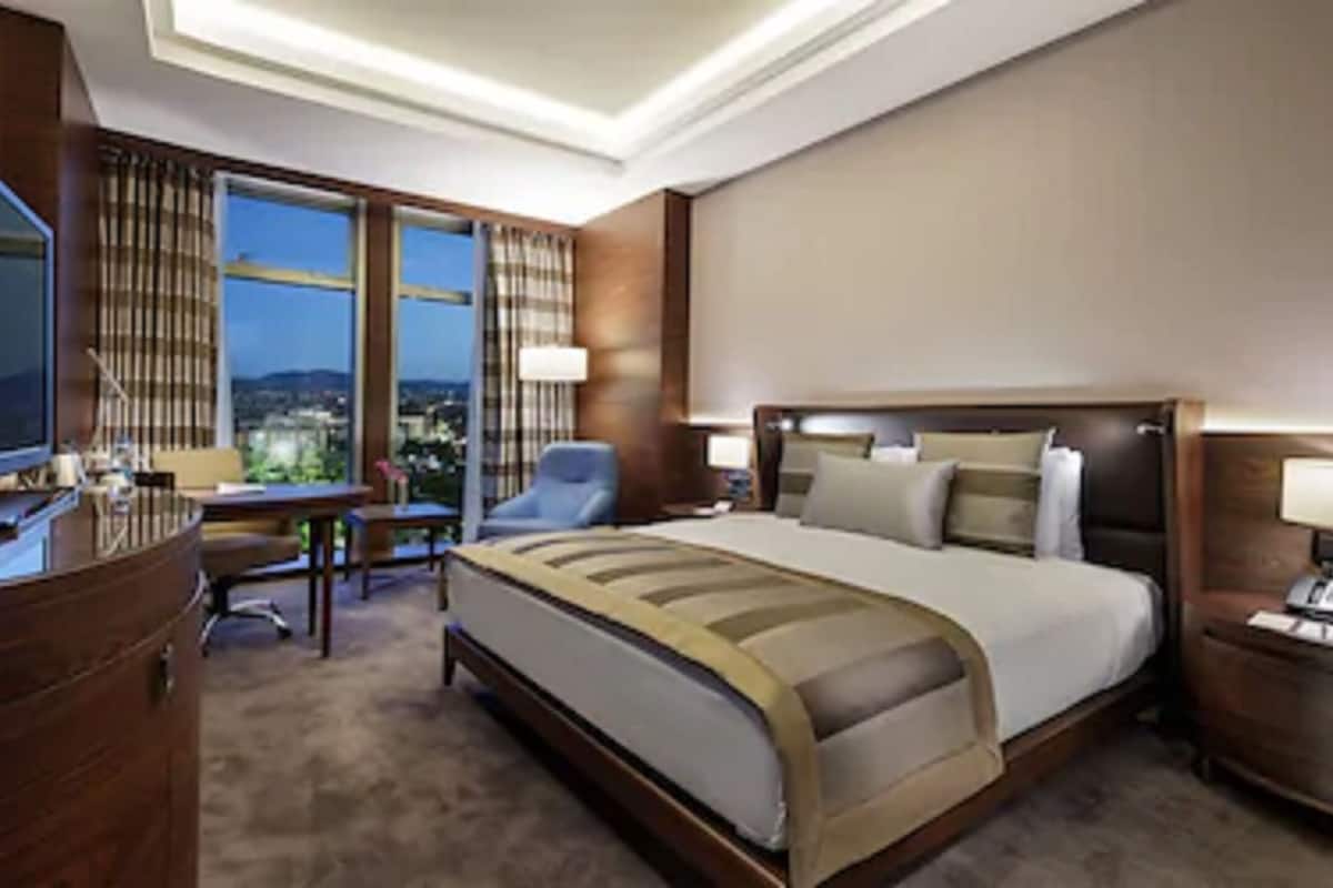 A hotel room with a large bed, a desk, a chair, a television, and a nightstand with a lamp.