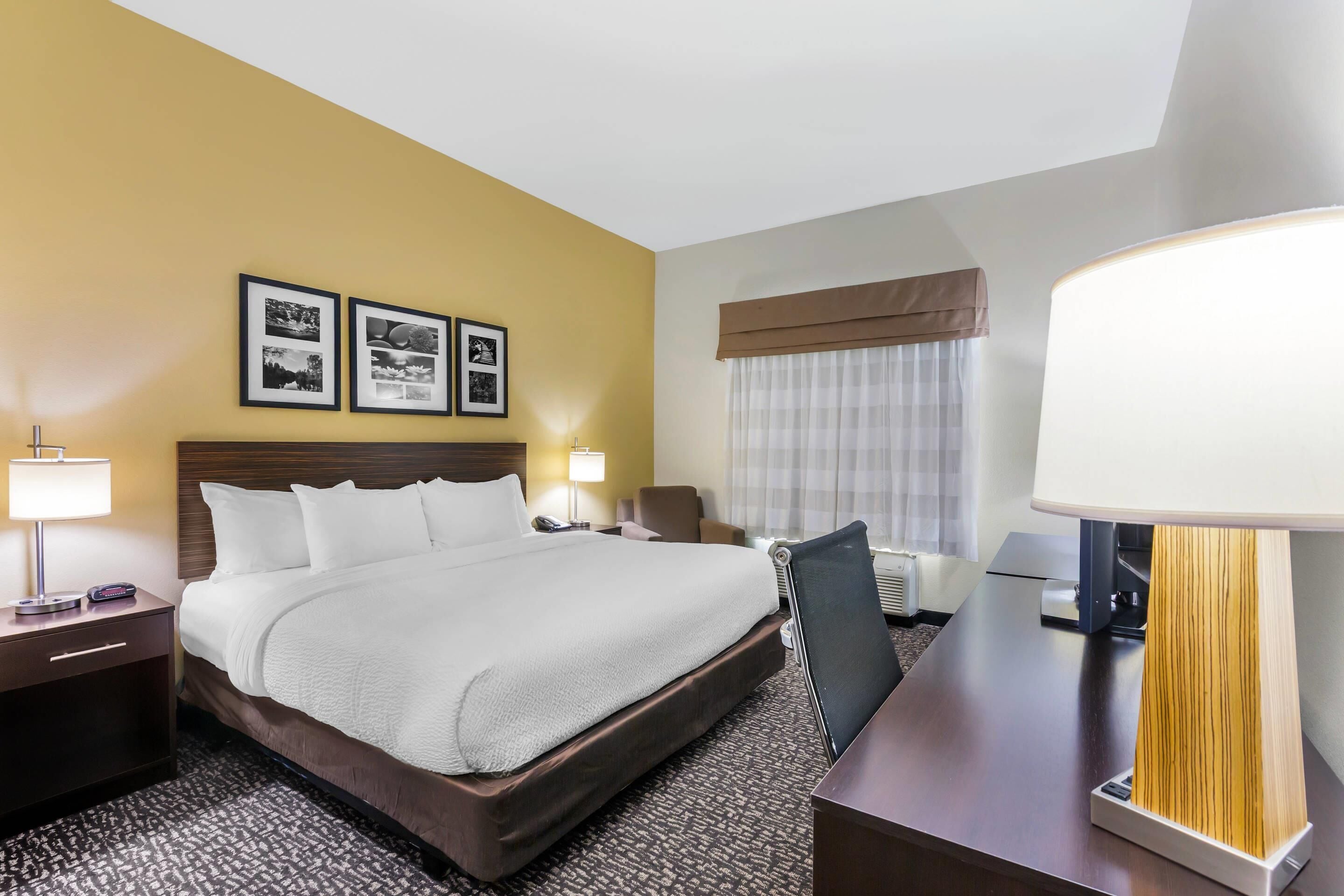 A hotel room with a large bed, two bedside lamps, a desk with a lamp, a chair, and a window with curtains.