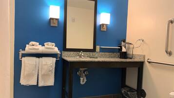 Un baño con pared azul, un lavabo con encimera de granito, un espejo, dos luces empotradas en la pared, un perchero con toallas, un cesto de basura y un pasamanos.