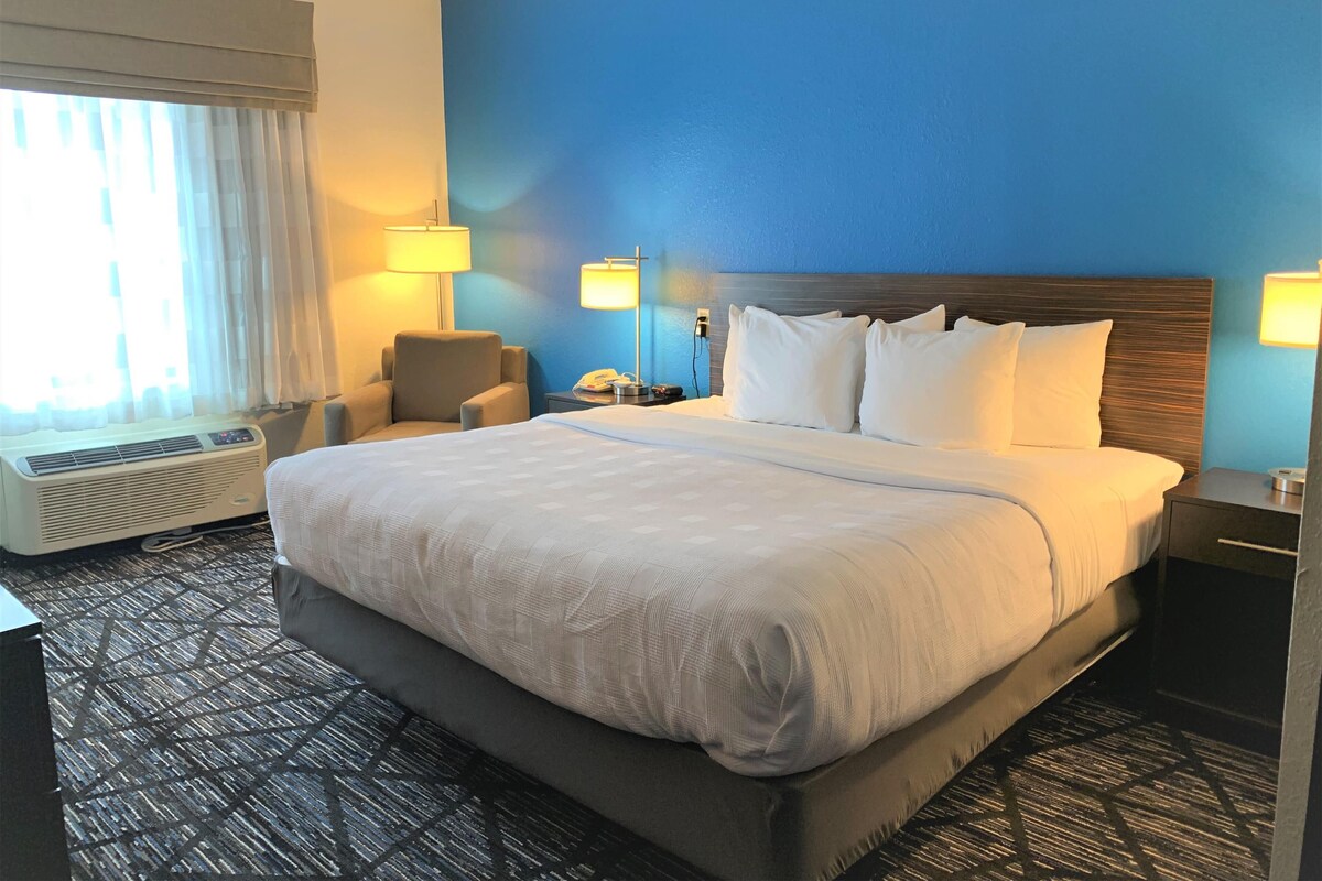 A hotel room with a large bed, two bedside lamps, a chair, a desk, and an air conditioning unit.