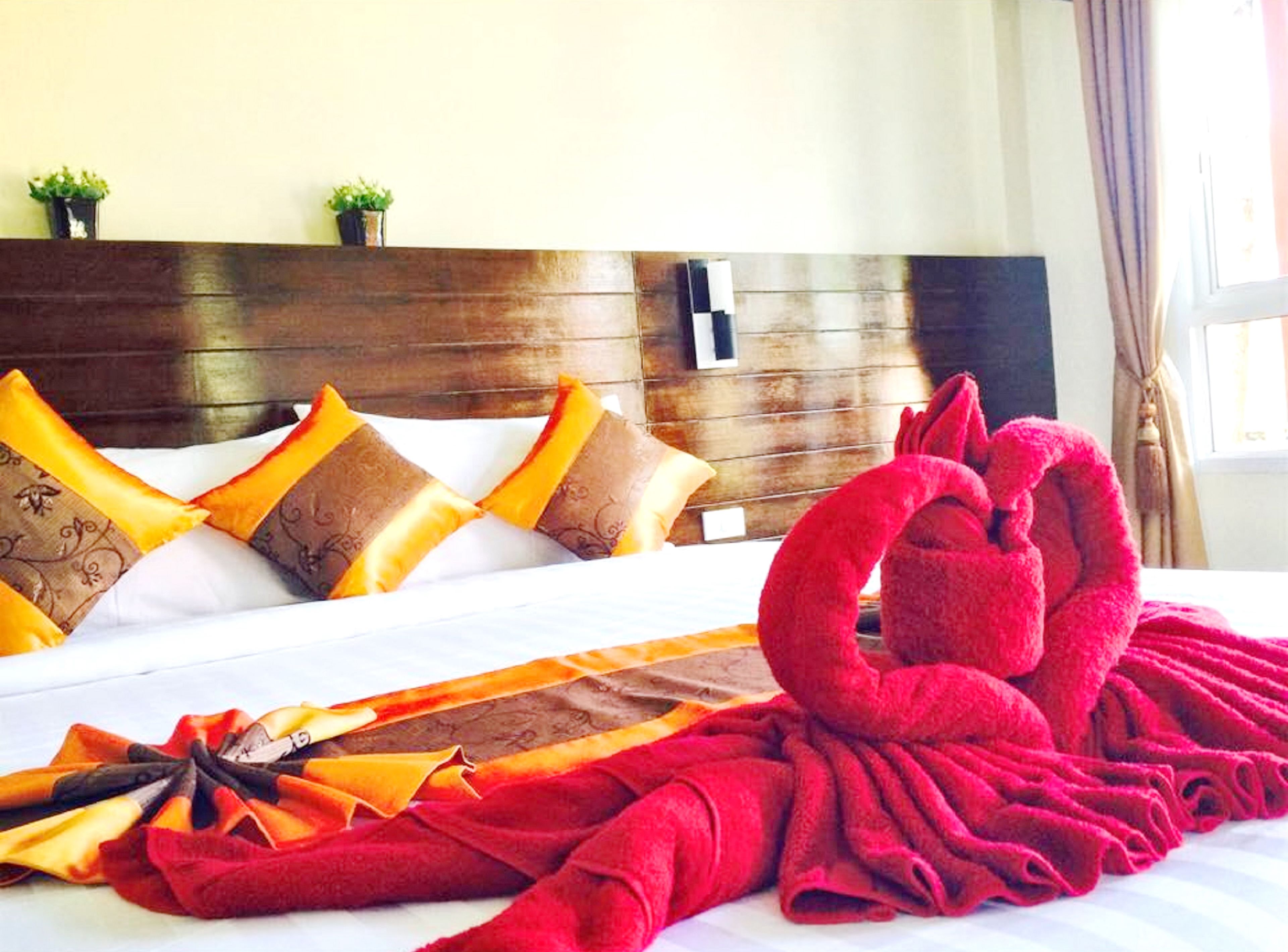 A hotel room with a wooden headboard, a bed with patterned pillows, and a red towel arranged in a swan shape.