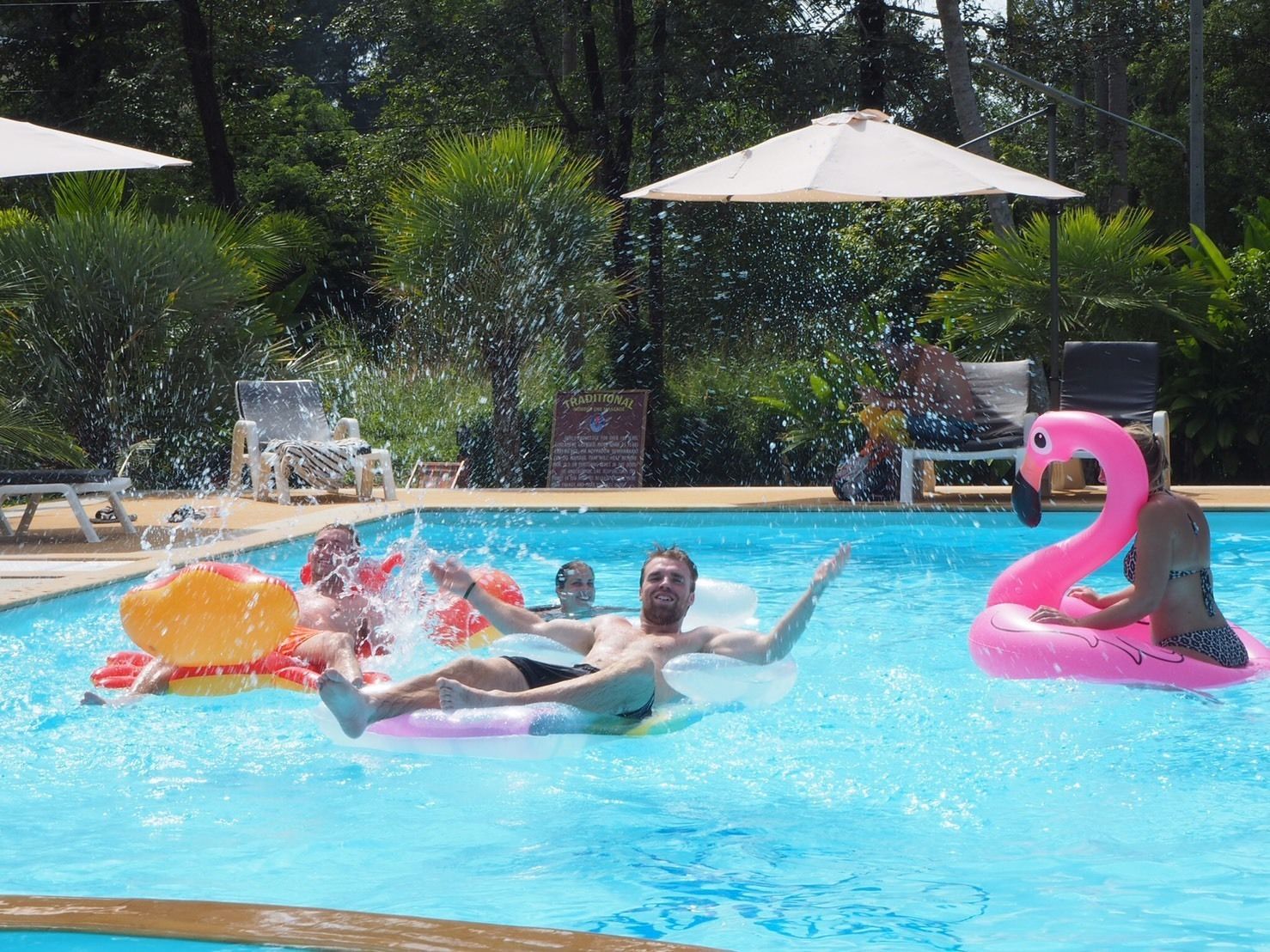 Una piscina amb persones sobre joguets flotants, envoltada de gandules i paraigudes.