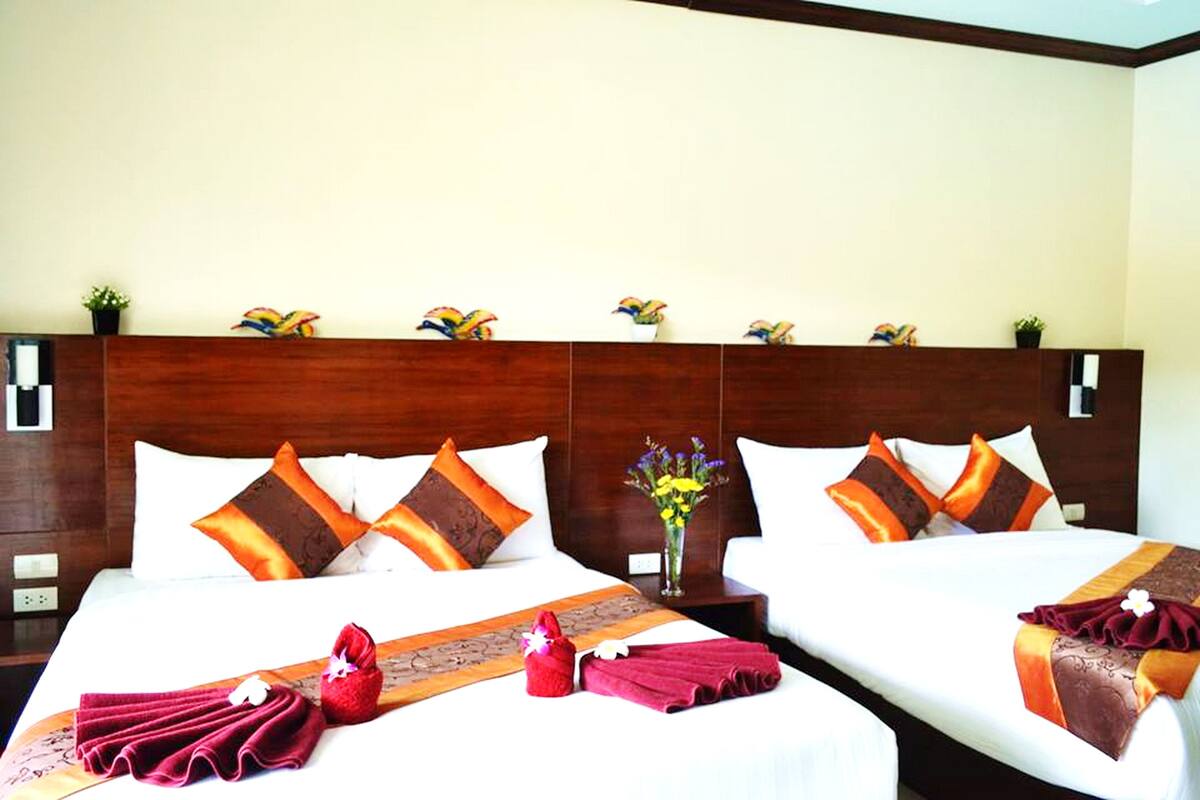 Une chambre d’hôtel avec deux lits, des têtes de lit en bois, du linge de lit blanc, des oreillers orange et marron, un vase avec des fleurs et une serviette rouge sur le lit.