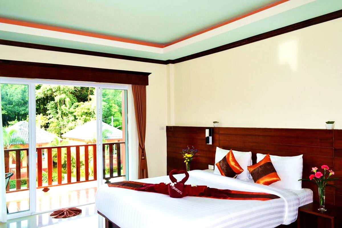 Une chambre d’hôtel avec un grand lit, une tête de lit en bois, une fenêtre donnant sur un paysage verdoyant et un balcon avec une balustrade.