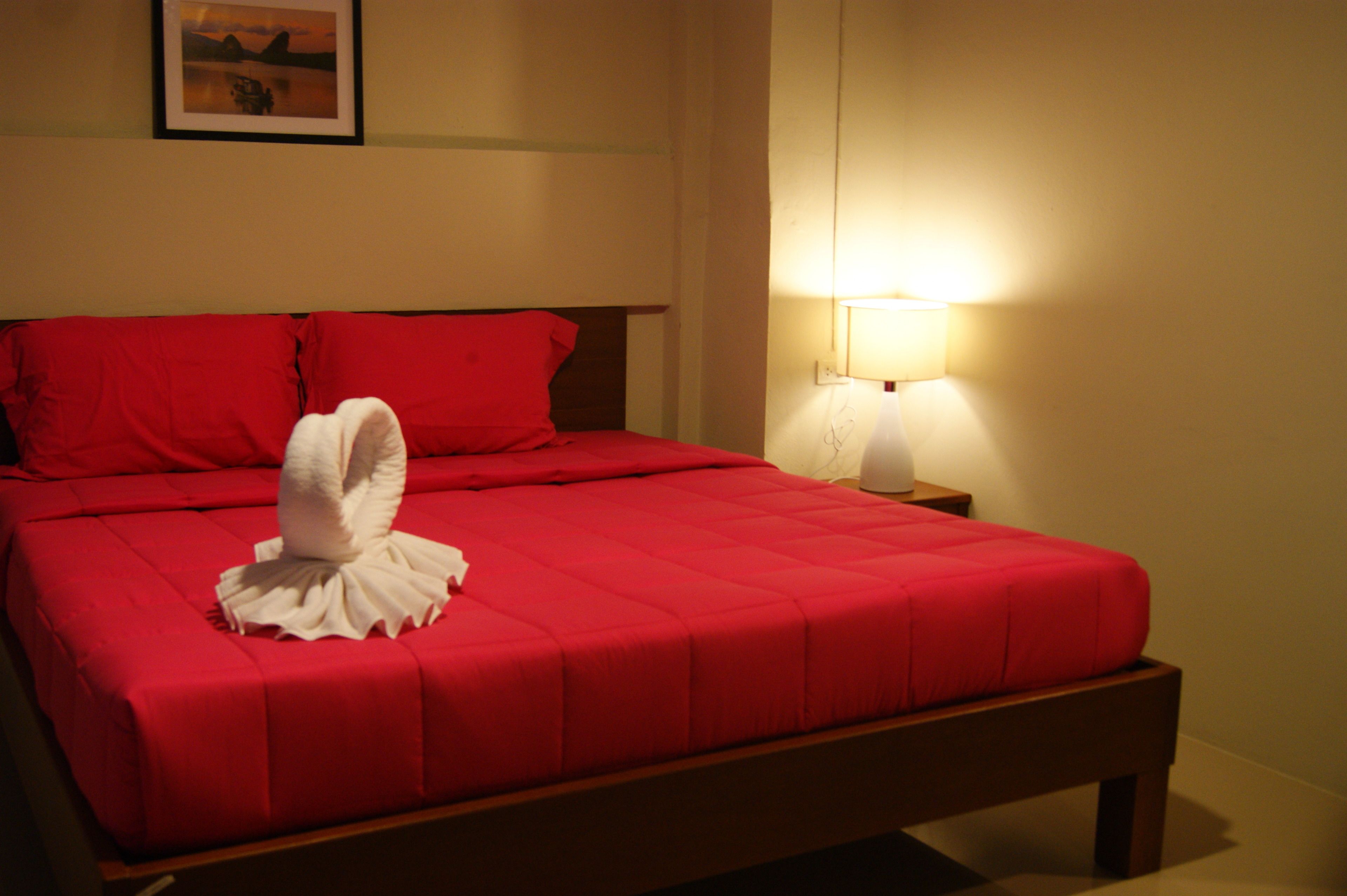 Un lit bien fait, avec une couverture rouge et des oreillers, une serviette blanche disposée en forme de cygne, et une lampe de chevet posée sur une table de nuit en bois.