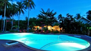 Ein Pool mit geschwungener Kante, umgeben von Palmen und einem Gebäude mit mehreren Fenstern und beleuchteter Fassade.